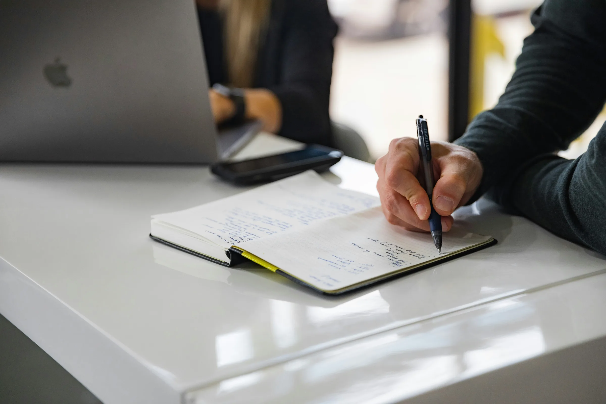 Main prenant des notes au stylo sur un carnet, ordinateur portable en arrière-plan
